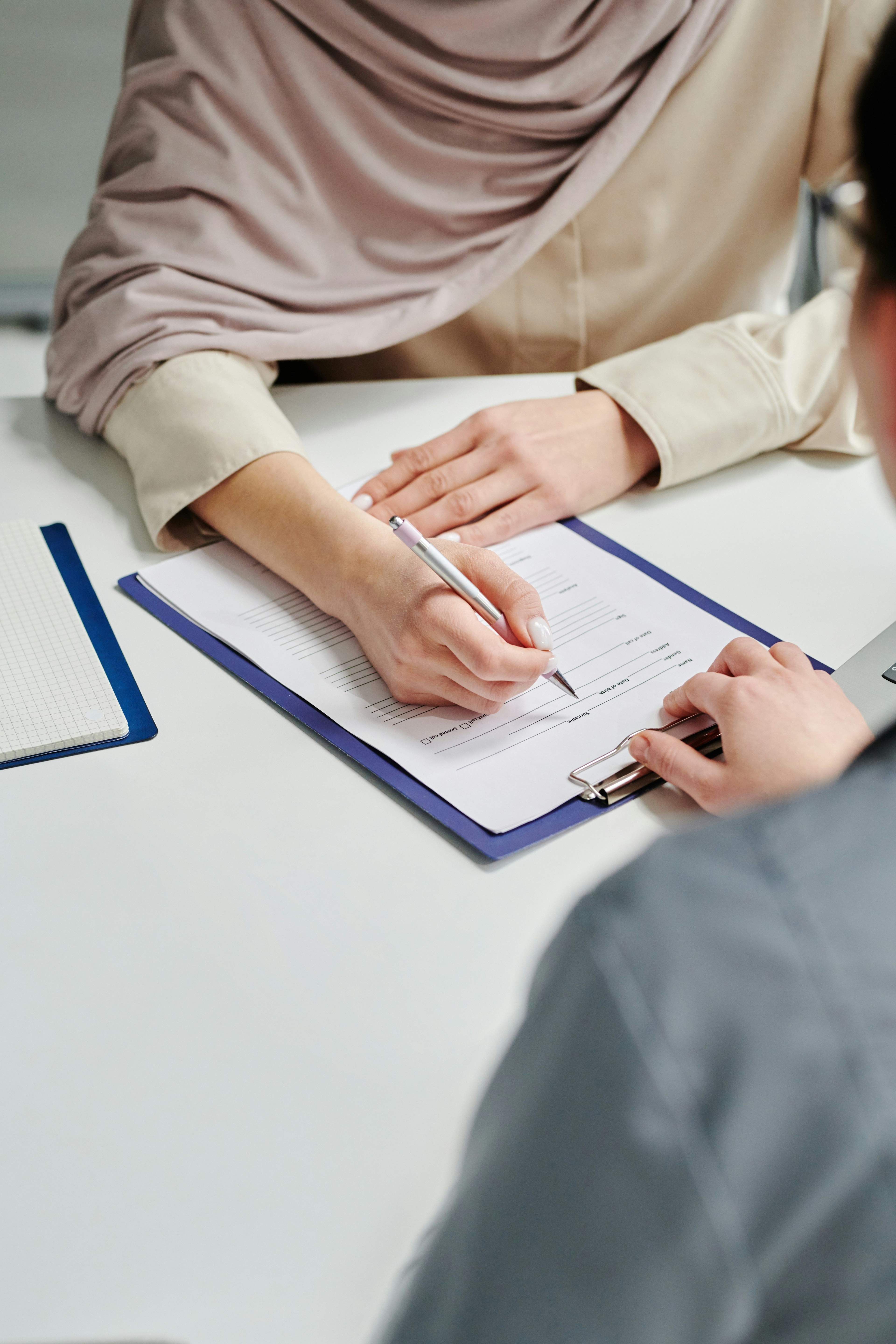 A person in a headscarf signs a document on a clipboard while another person is present.