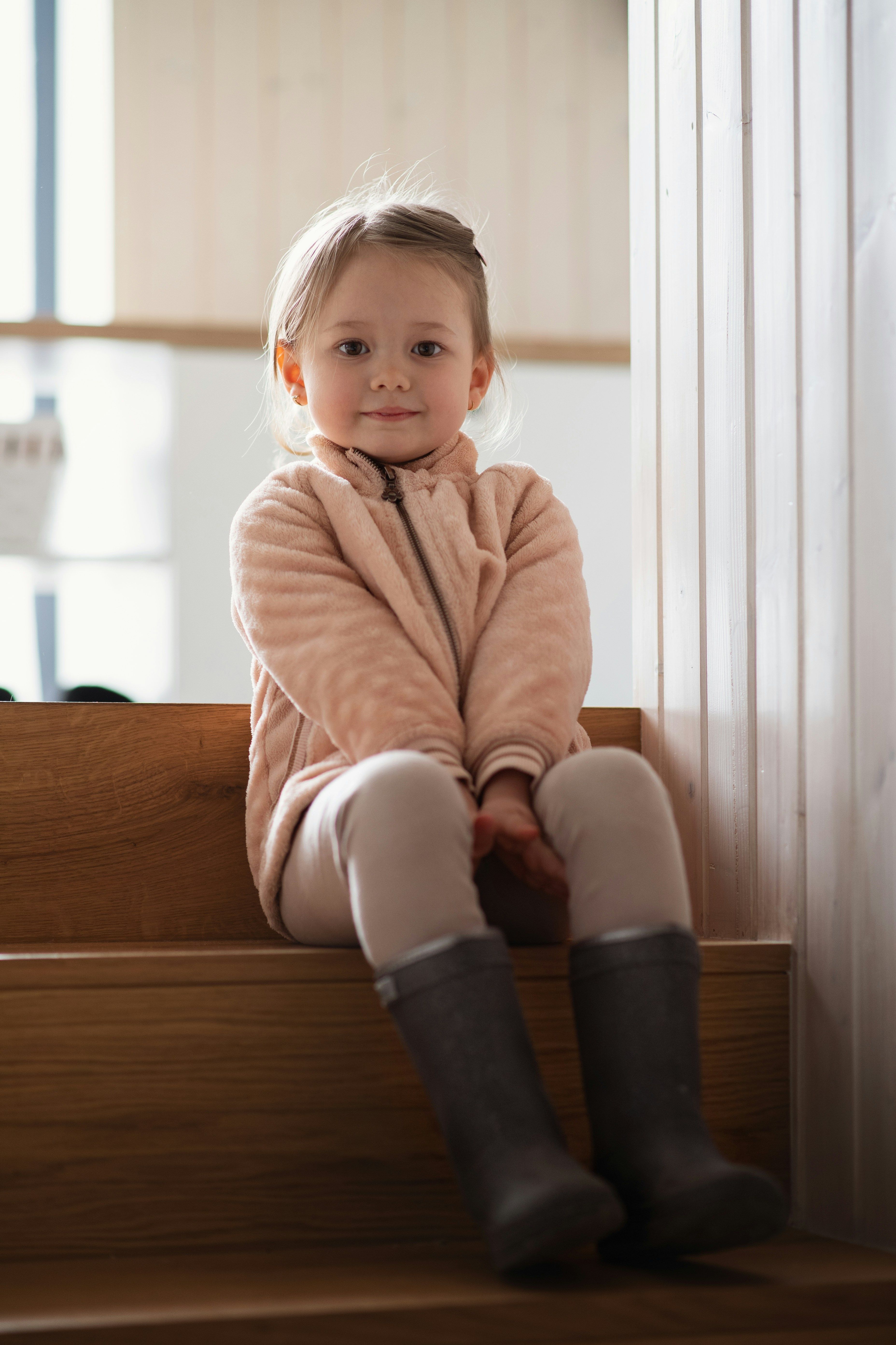 A young girl in a pink quilted jacket and grey boots sits on wooden stairs.