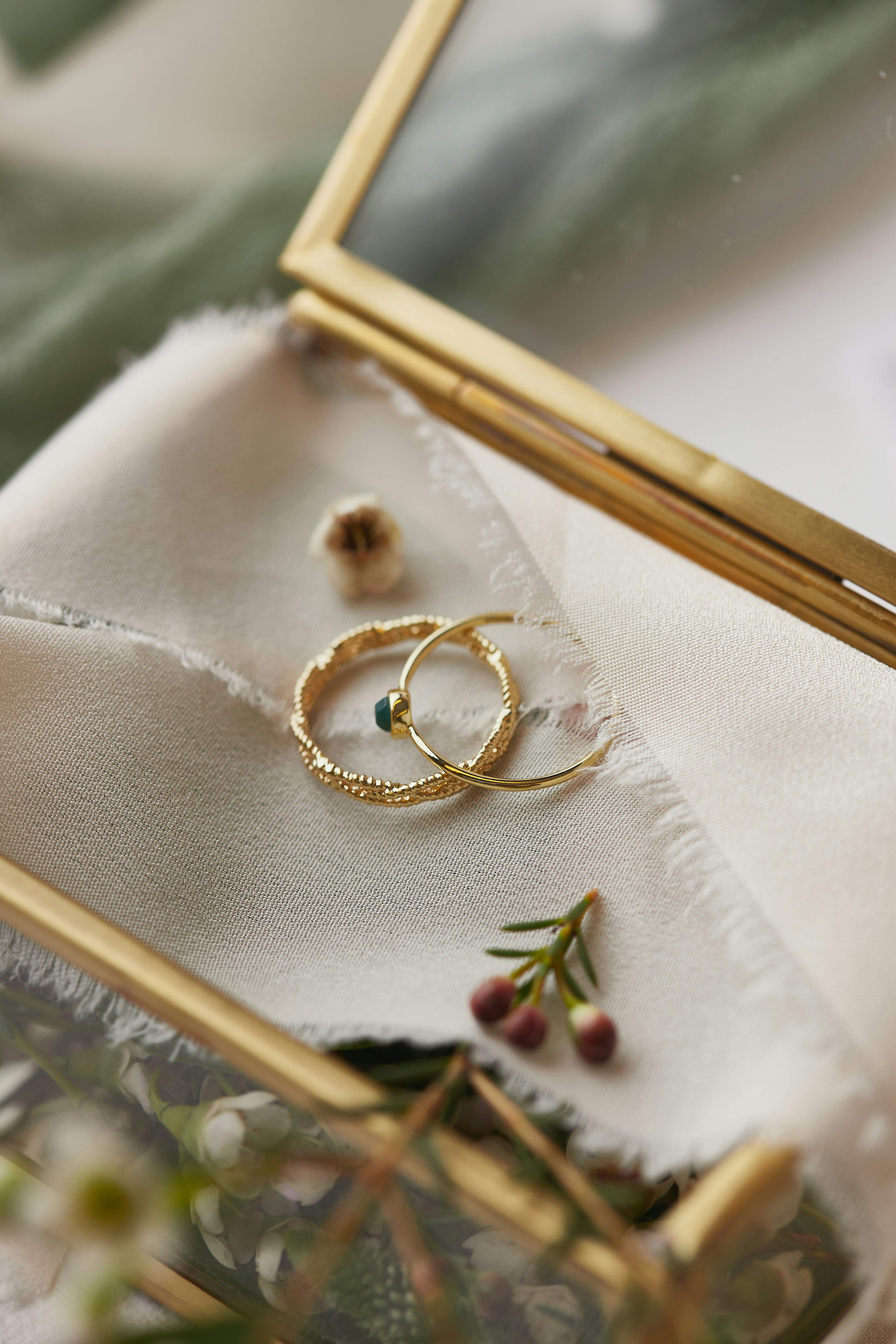 A delicate gold ring and earrings rest on a white fabric inside a golden glass box.