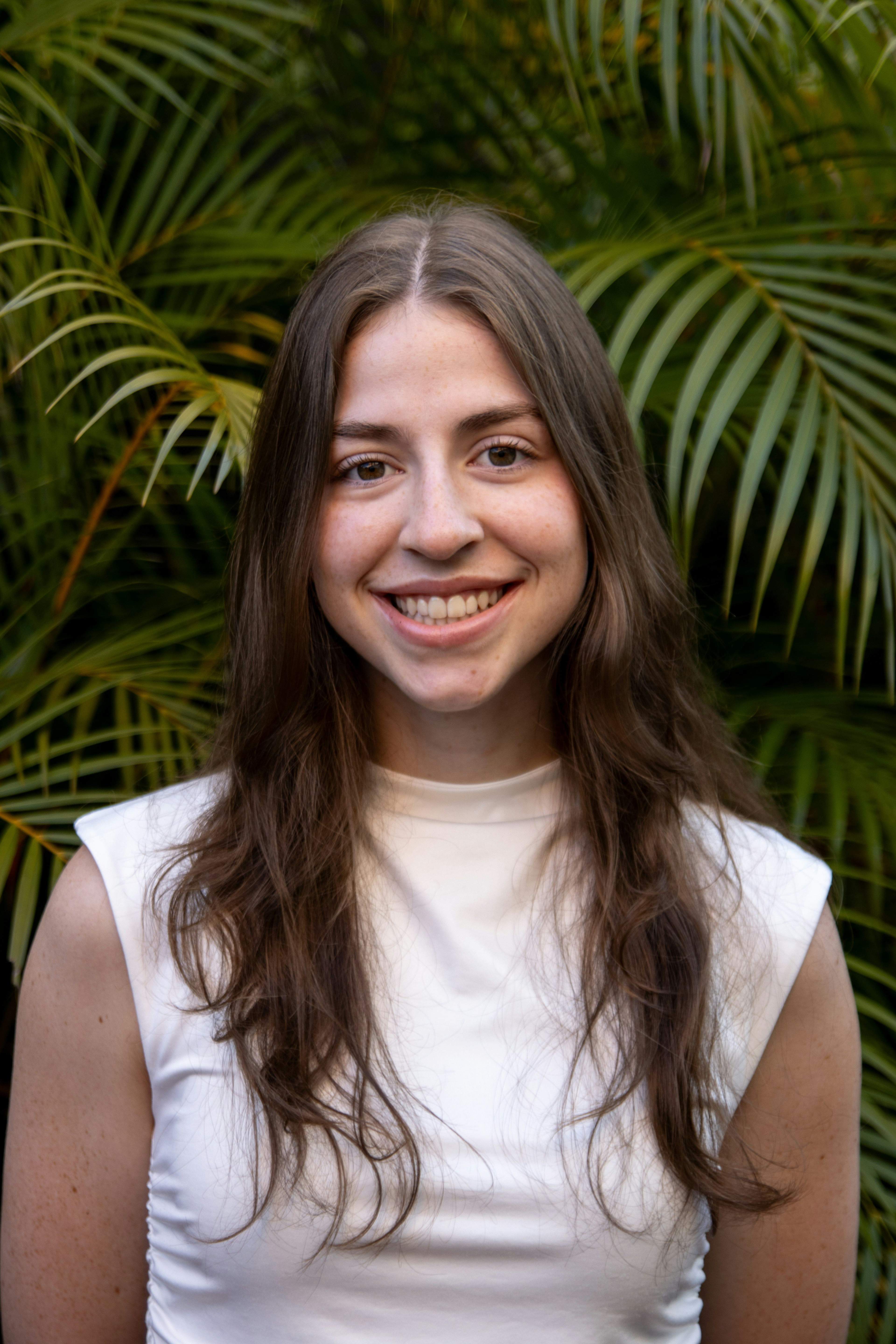 Smiling young woman with long brown hair wearing a white top, green plants in background.