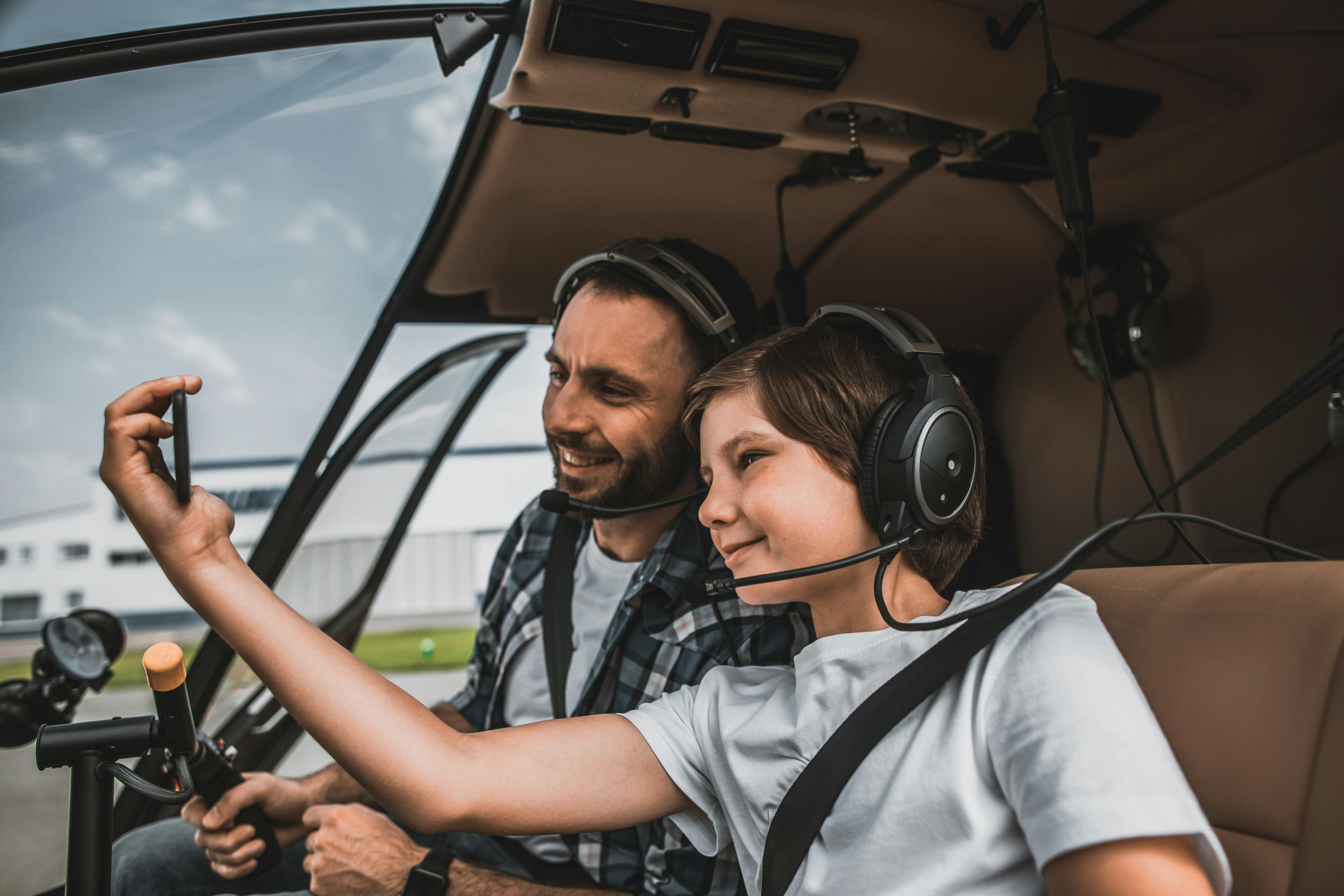 A smiling man and boy wear headsets, taking a selfie inside a helicopter.