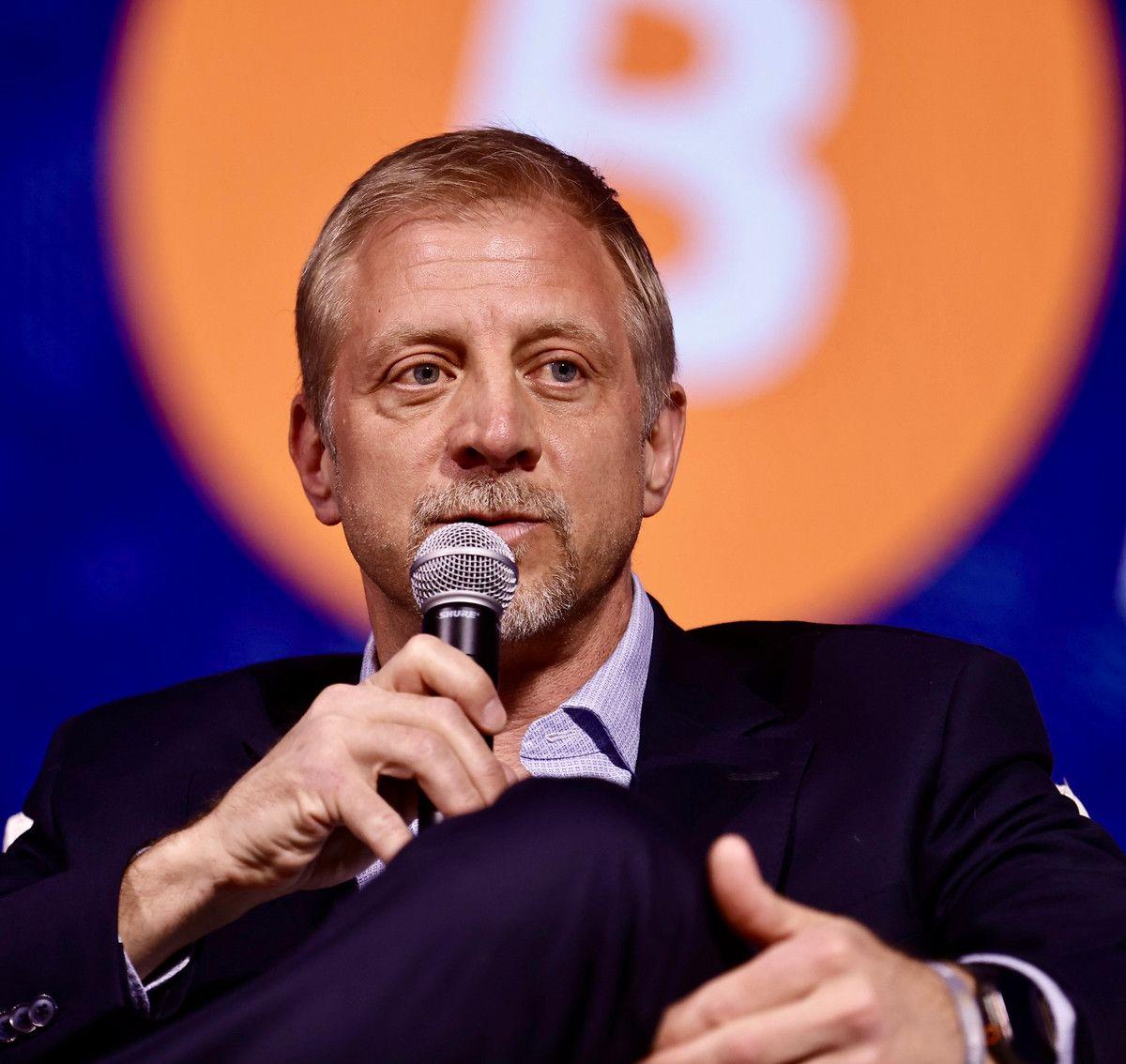 Man speaking into a microphone with a prominent Bitcoin logo in the background.
