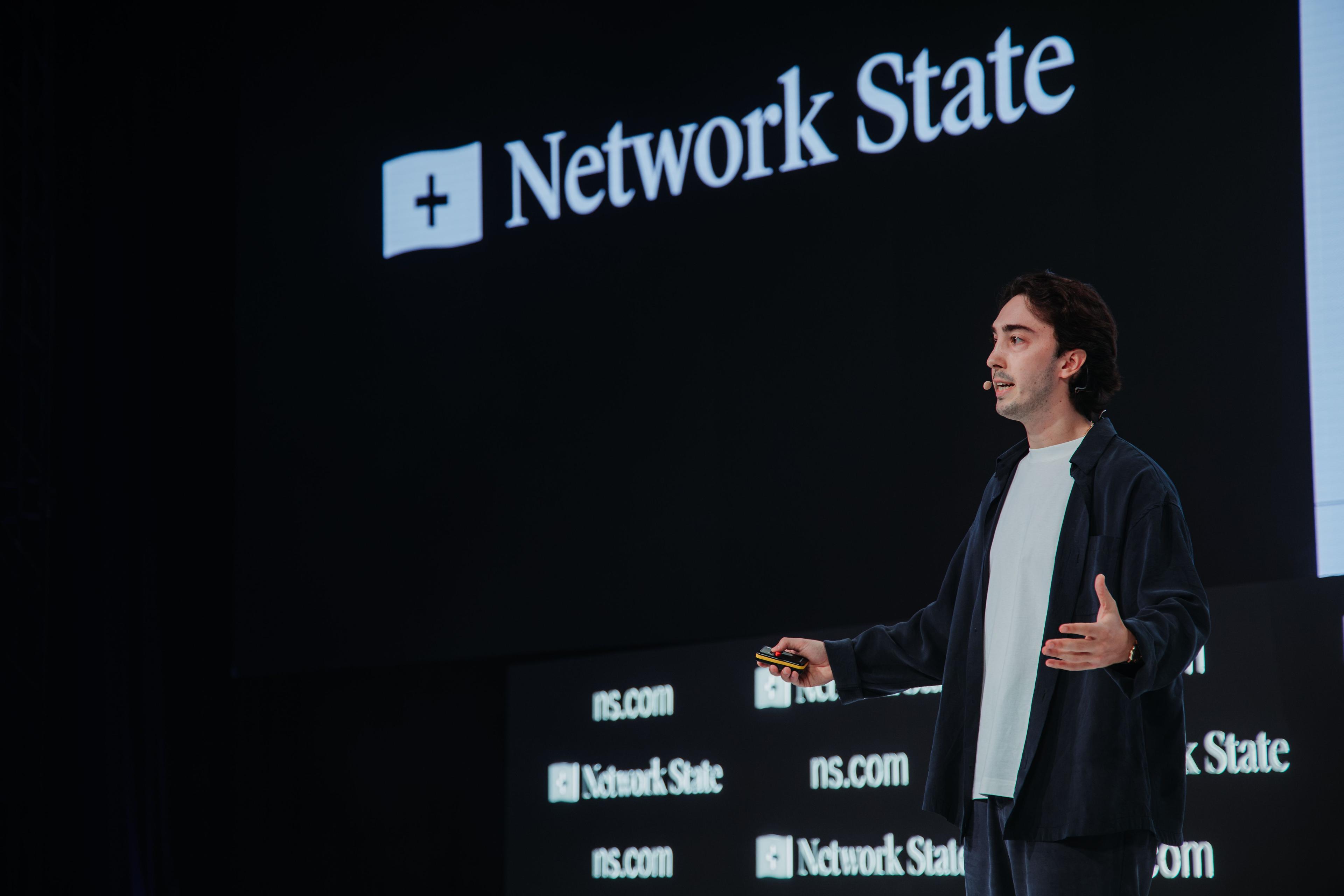 A man presents on stage in front of a screen displaying 'Network State'.