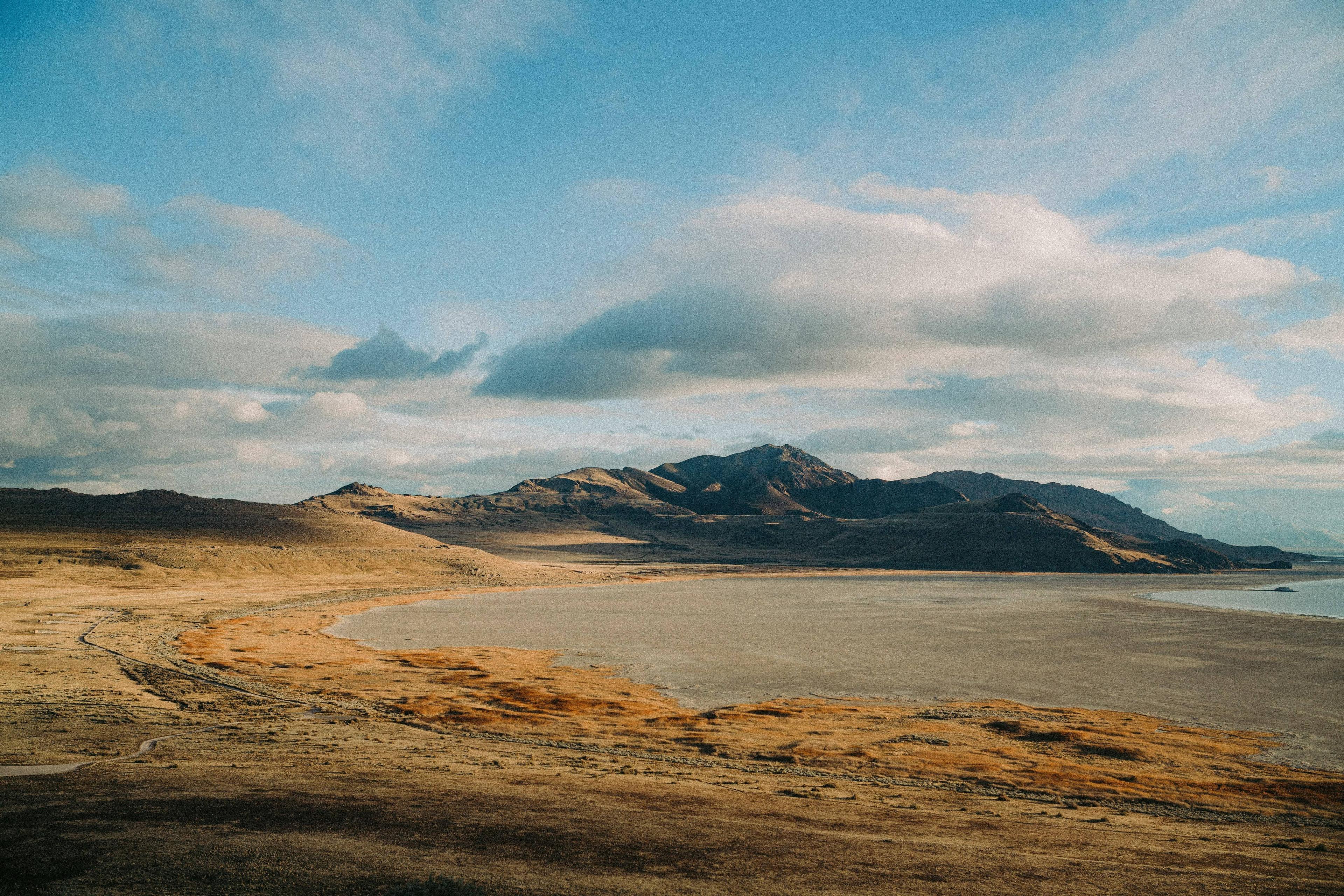 Arid landscape with golden hills, a dry lakebed, and distant mountains under cloudy sky.