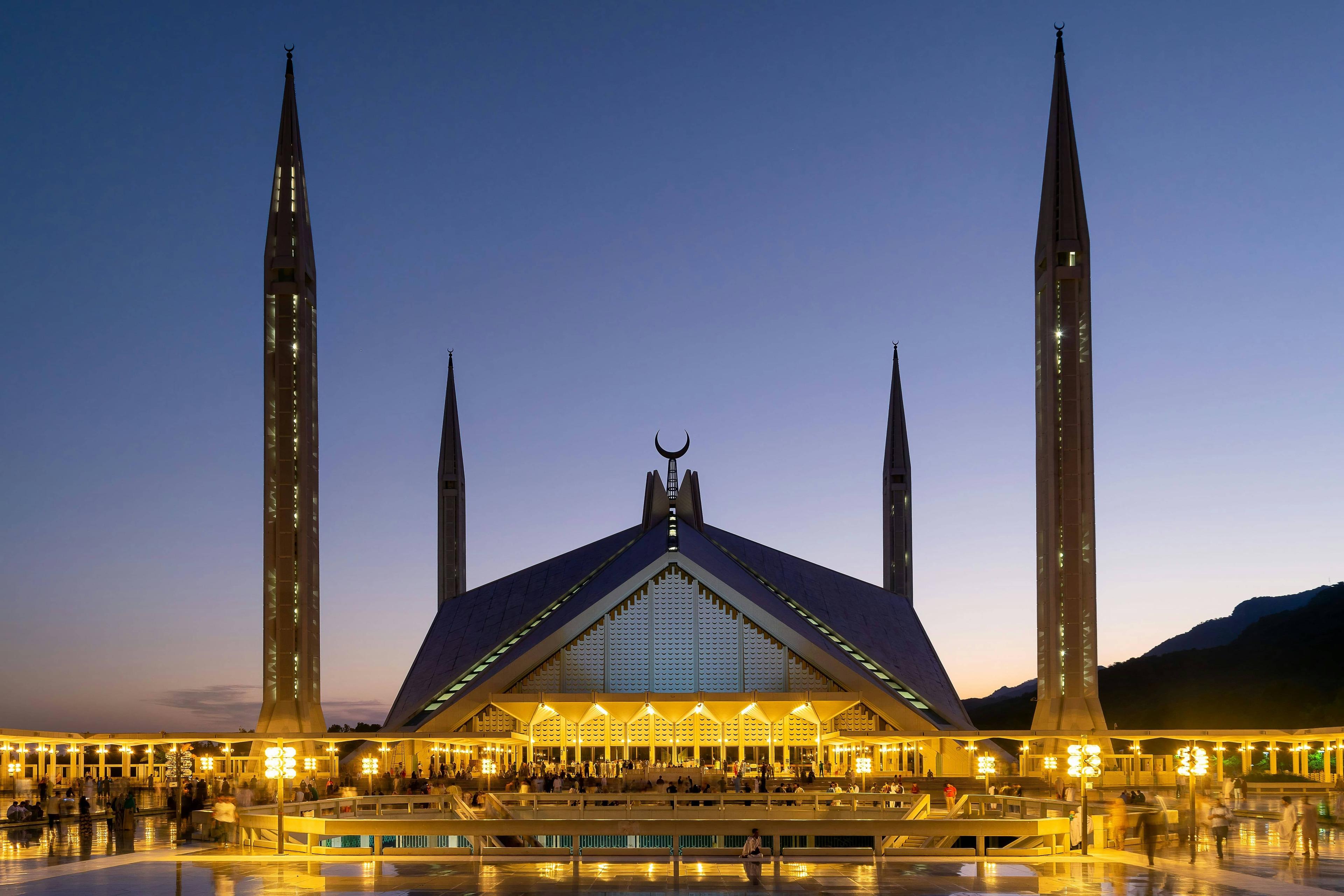 Faisal Mosque at night, illuminated, with four tall minarets against a blue sky.