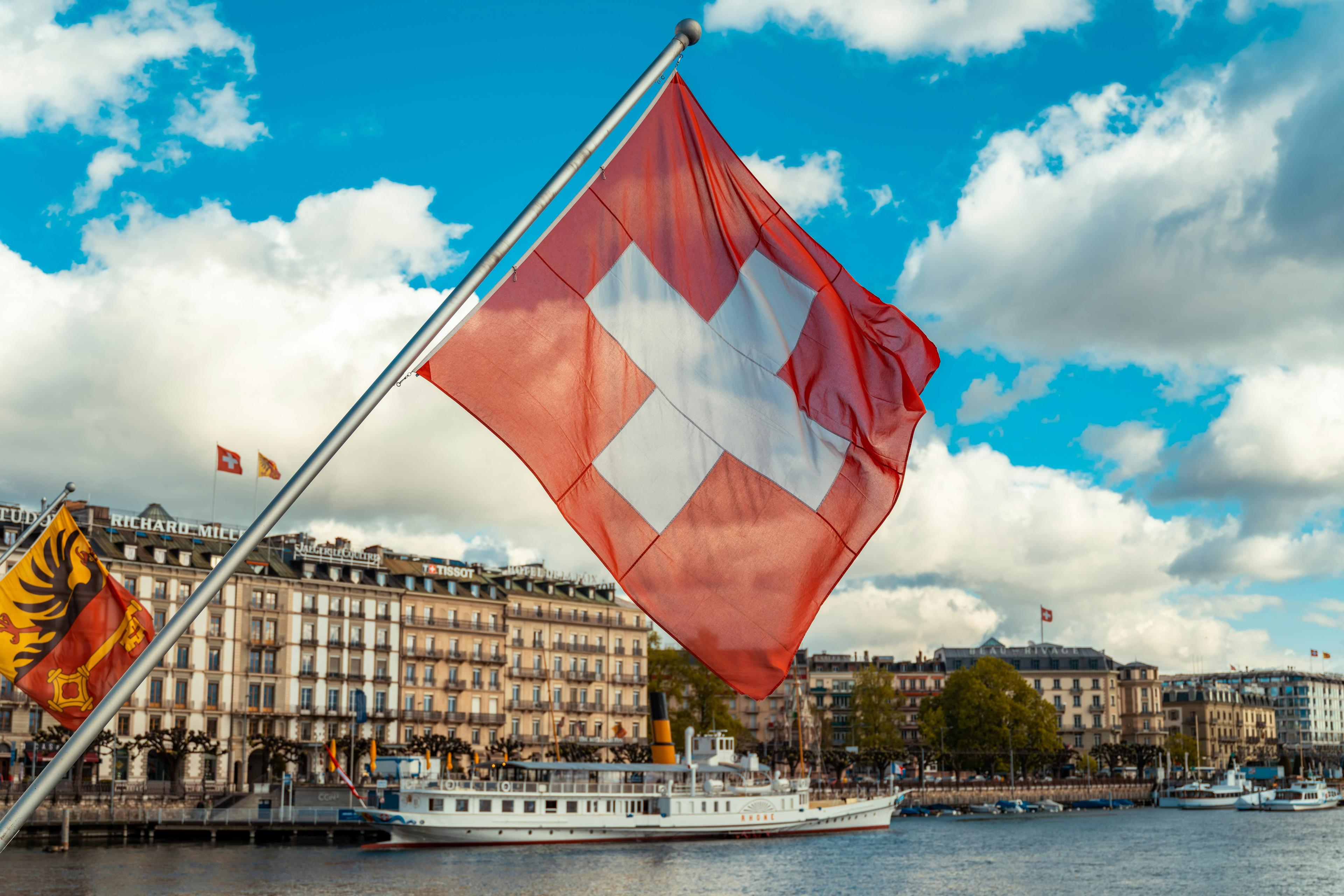Swiss flag waving in front of a European city skyline with a lake and boat.