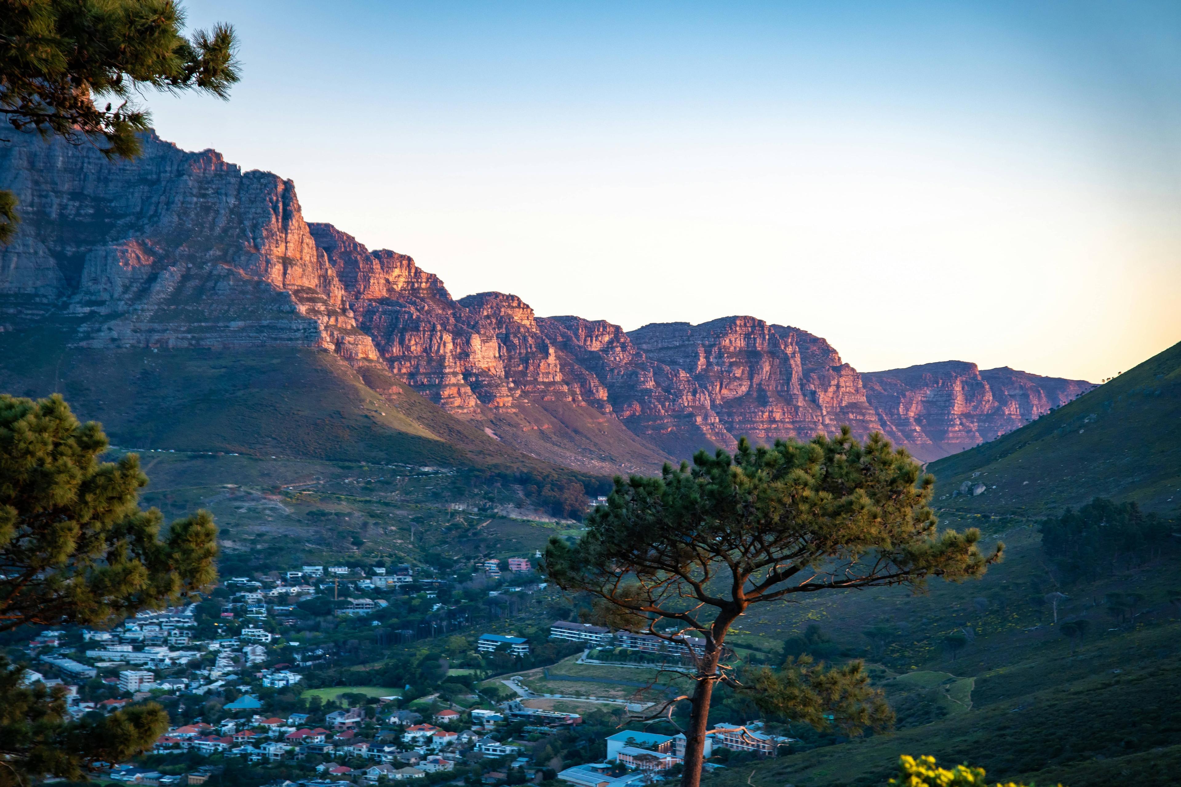 Golden sunlight illuminates rugged mountains overlooking a valley town with a prominent foreground tree.