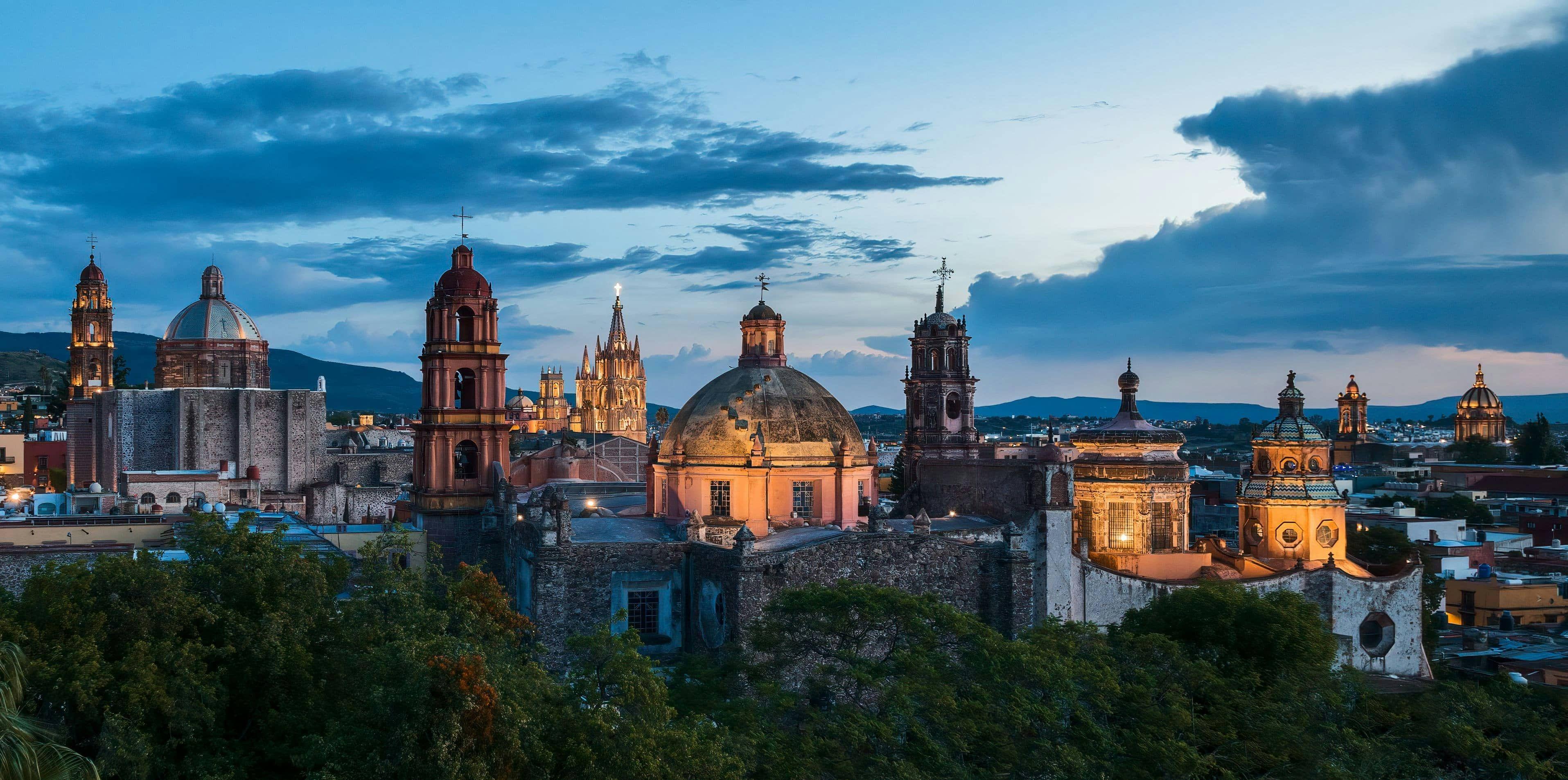 Historic city skyline at dusk with illuminated domes and spires against a cloudy sky.