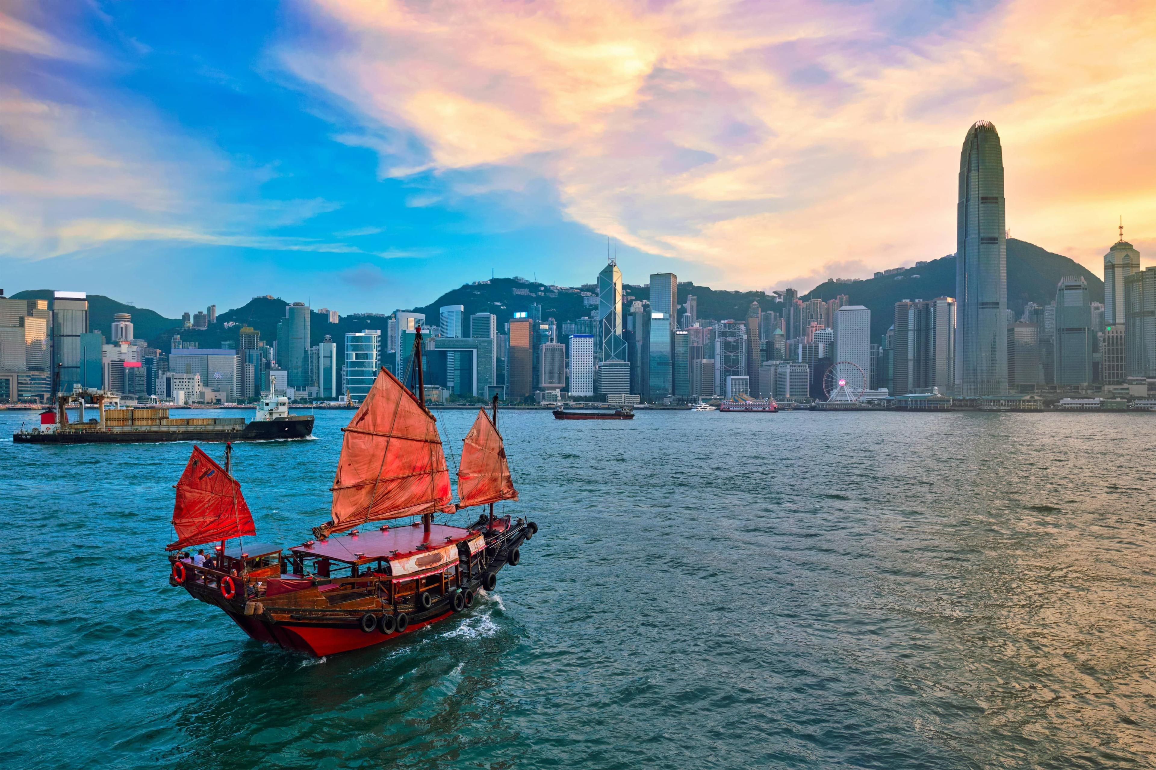 Traditional red-sailed junk boat sailing past a vibrant Hong Kong city skyline at sunset.