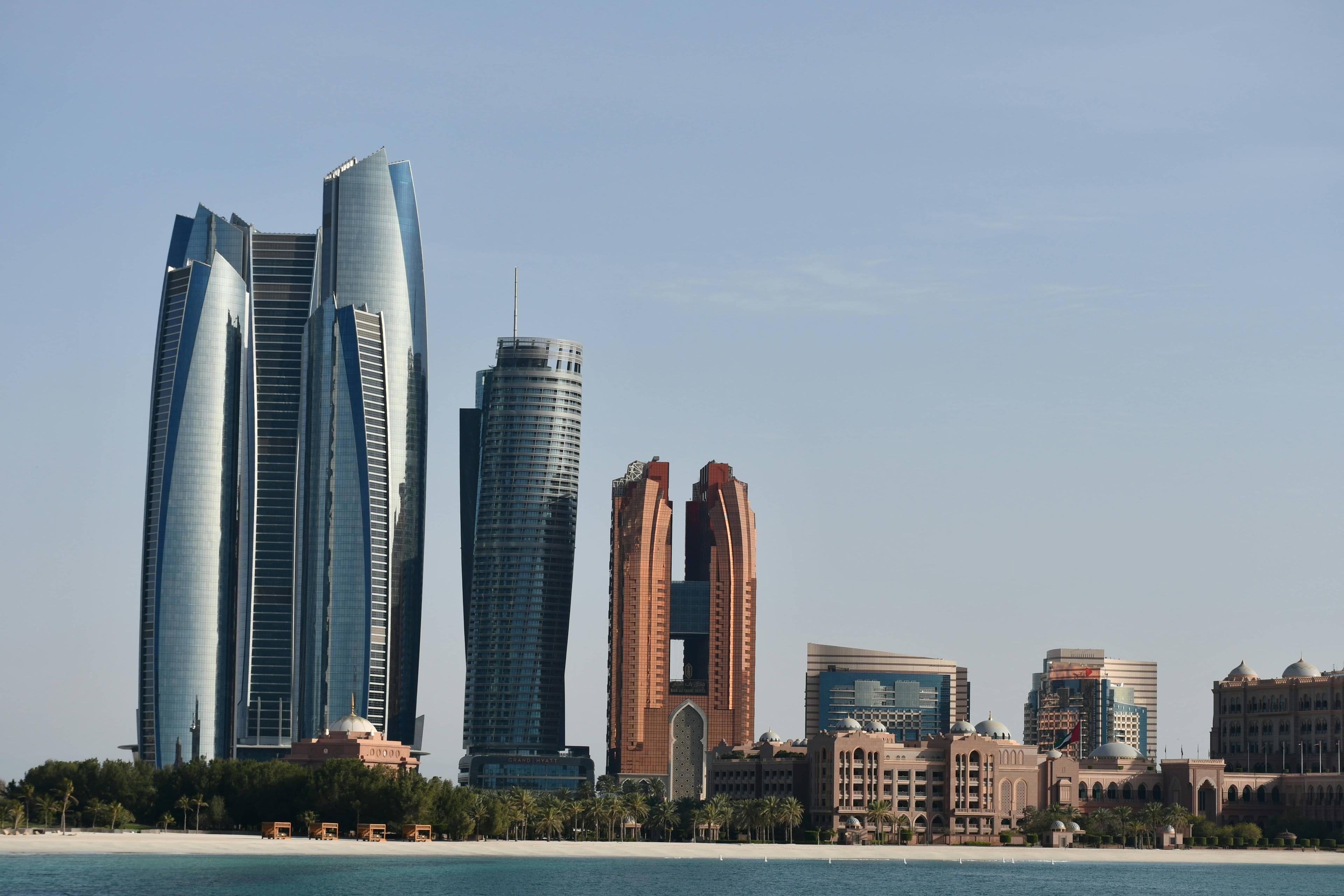 Futuristic skyscrapers in Abu Dhabi rise along the coastline against a clear blue sky.
