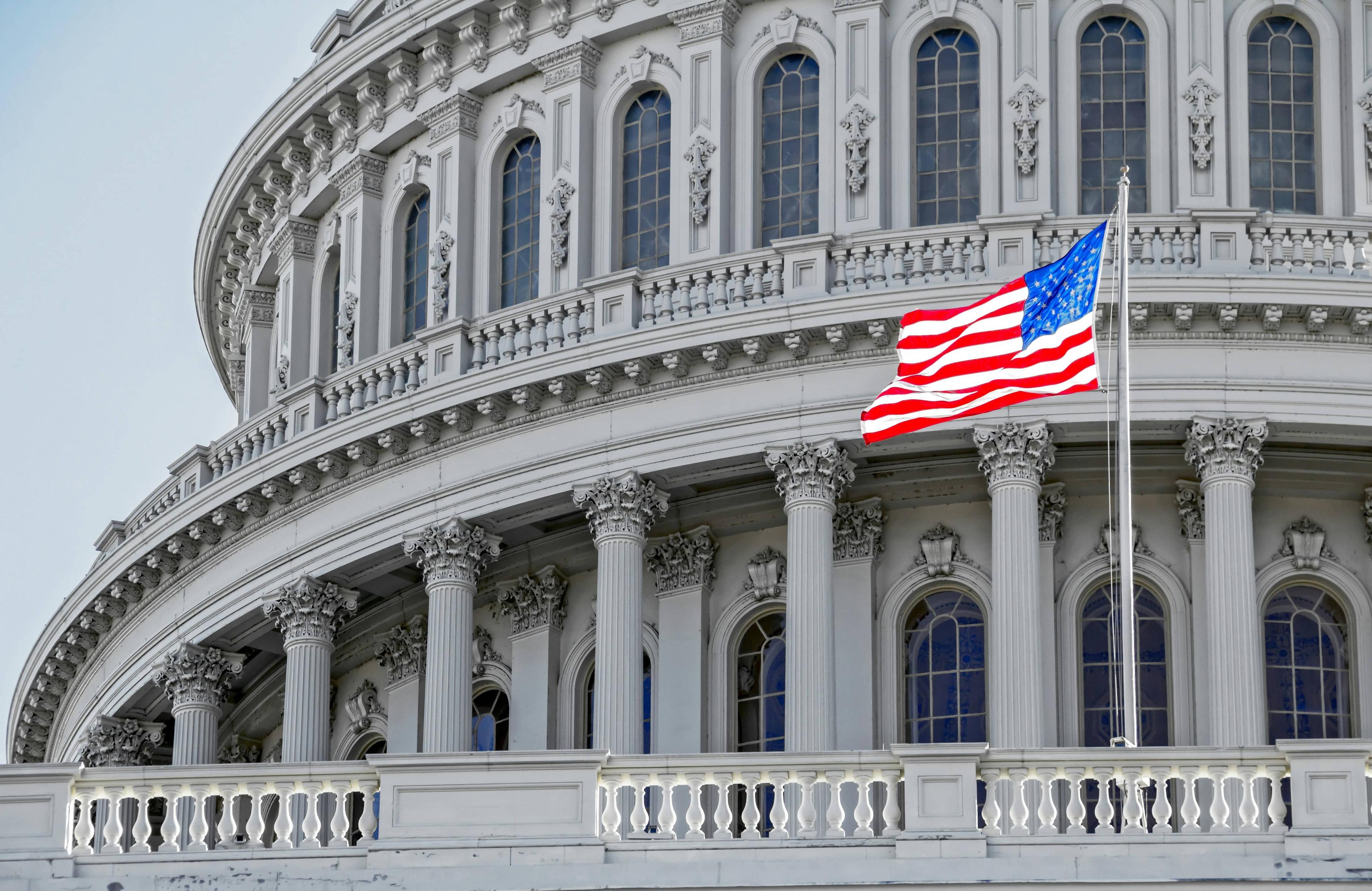 Close-up of the US Capitol Building dome, white columns, and an American flag flying.