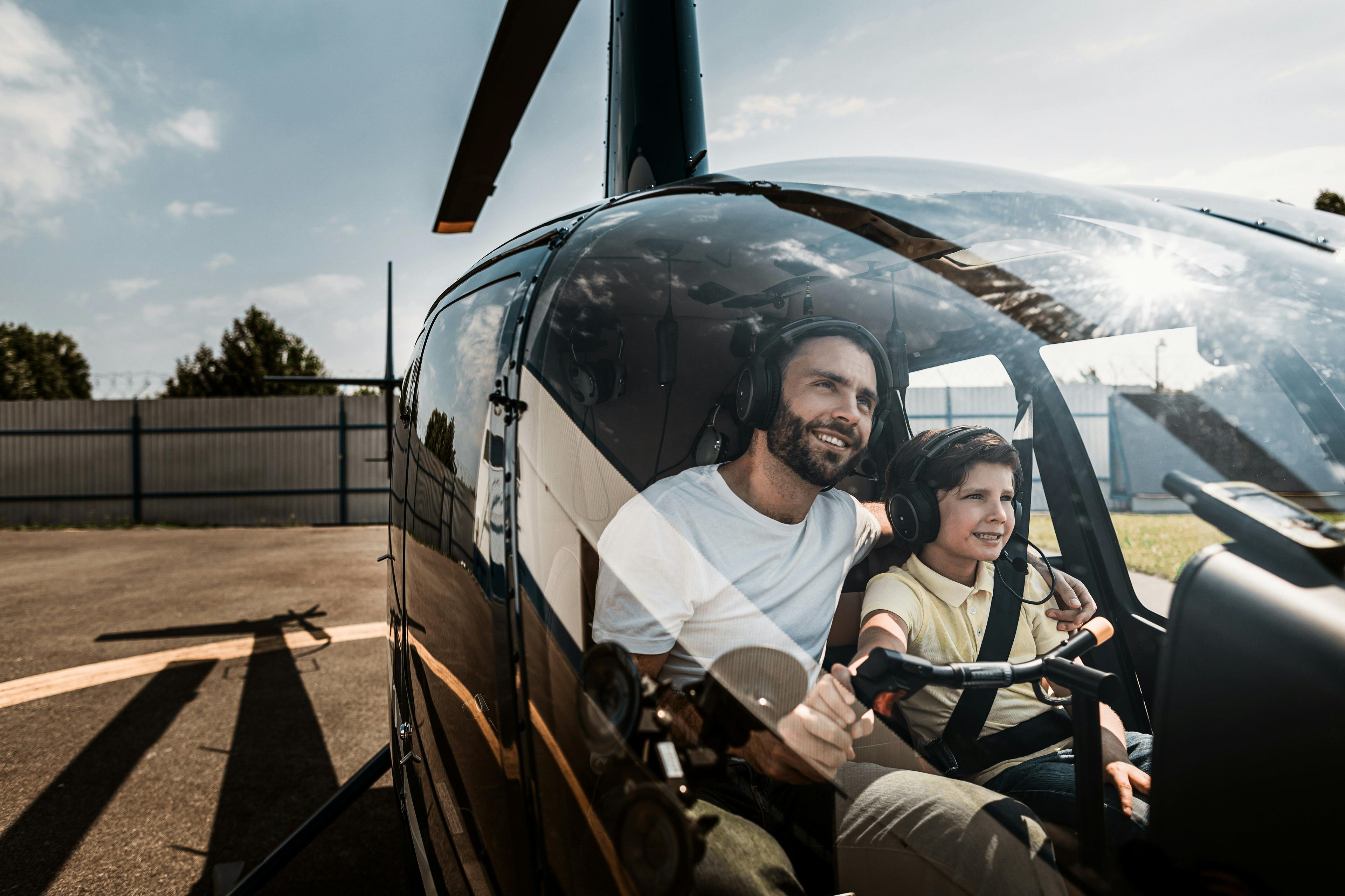 Smiling man and child sitting together in a black helicopter cockpit with headphones on.