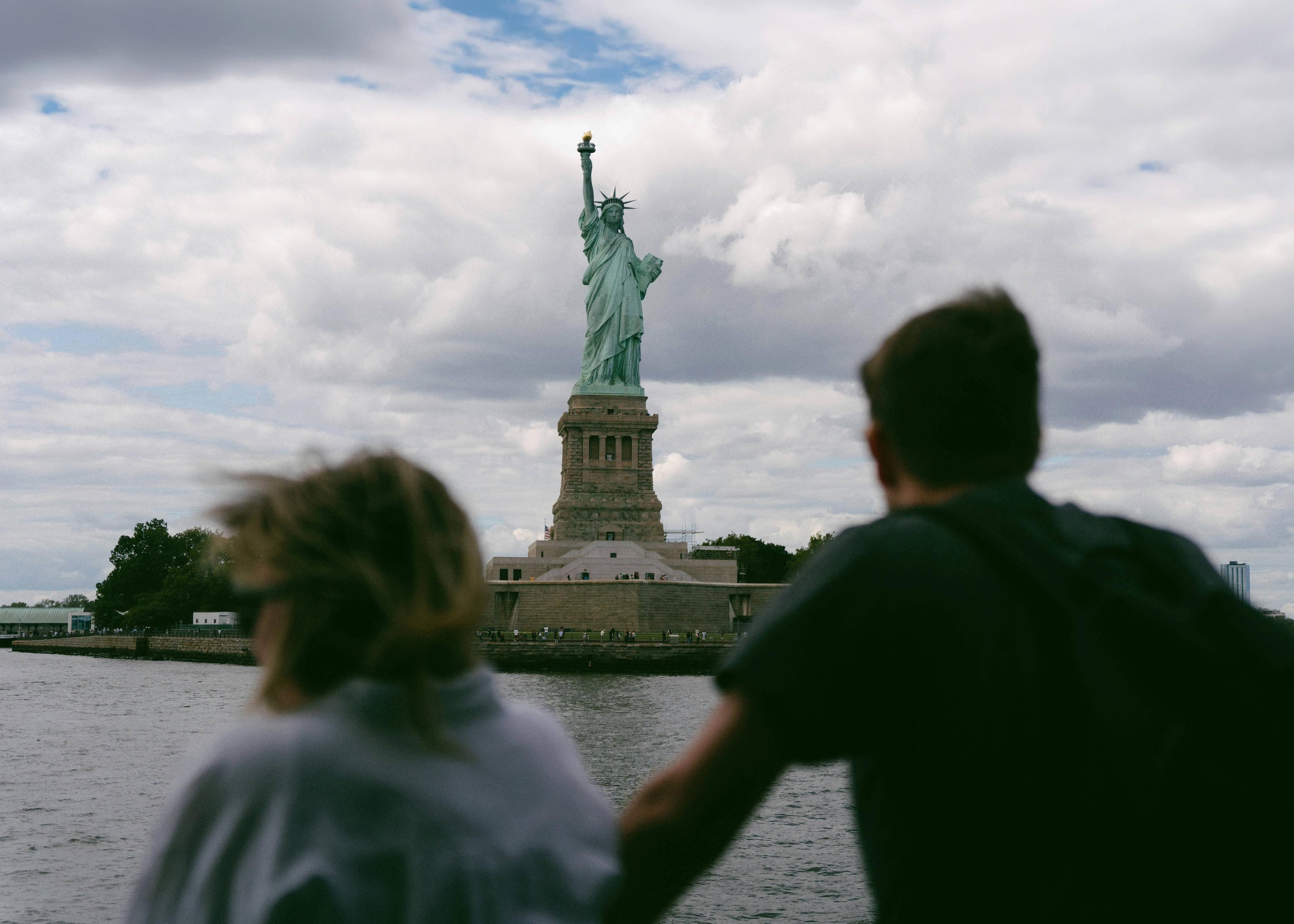 Two people on a boat gaze at the Statue of Liberty under a cloudy sky.
