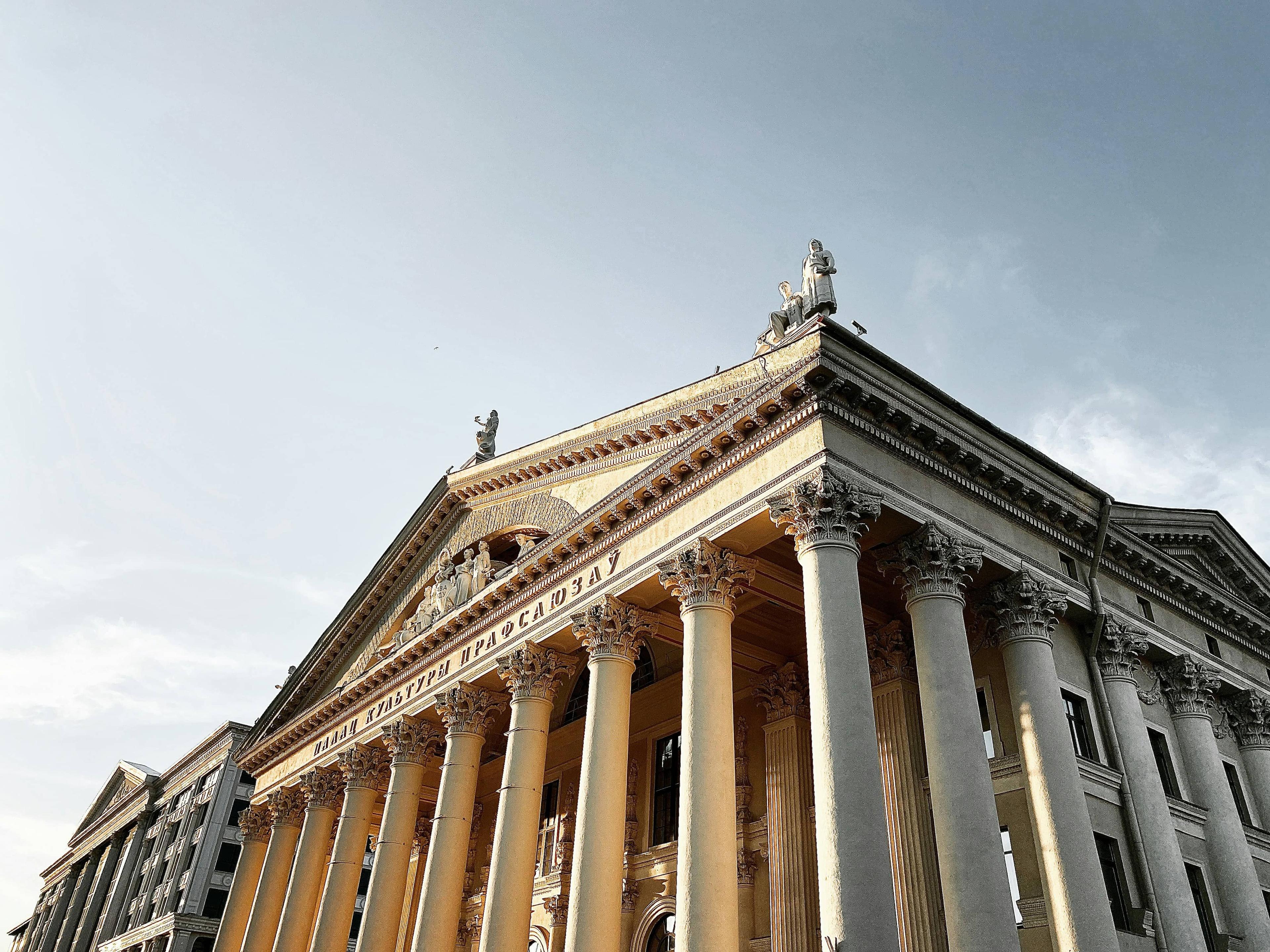 Grand classical building with tall columns, a pediment, and rooftop statues against a clear sky.