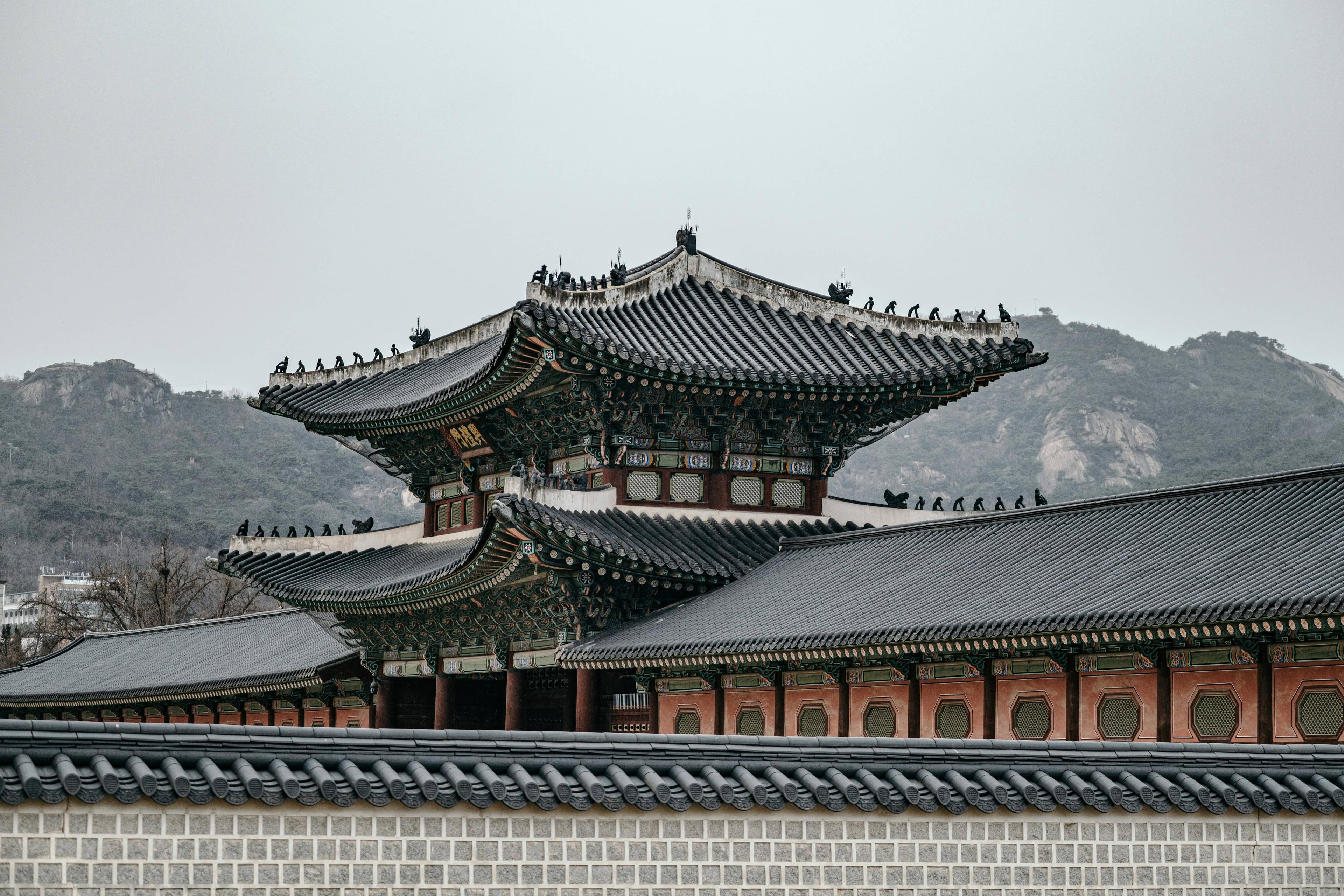 Traditional Korean palace with elaborate layered roof and mountains under a cloudy sky.