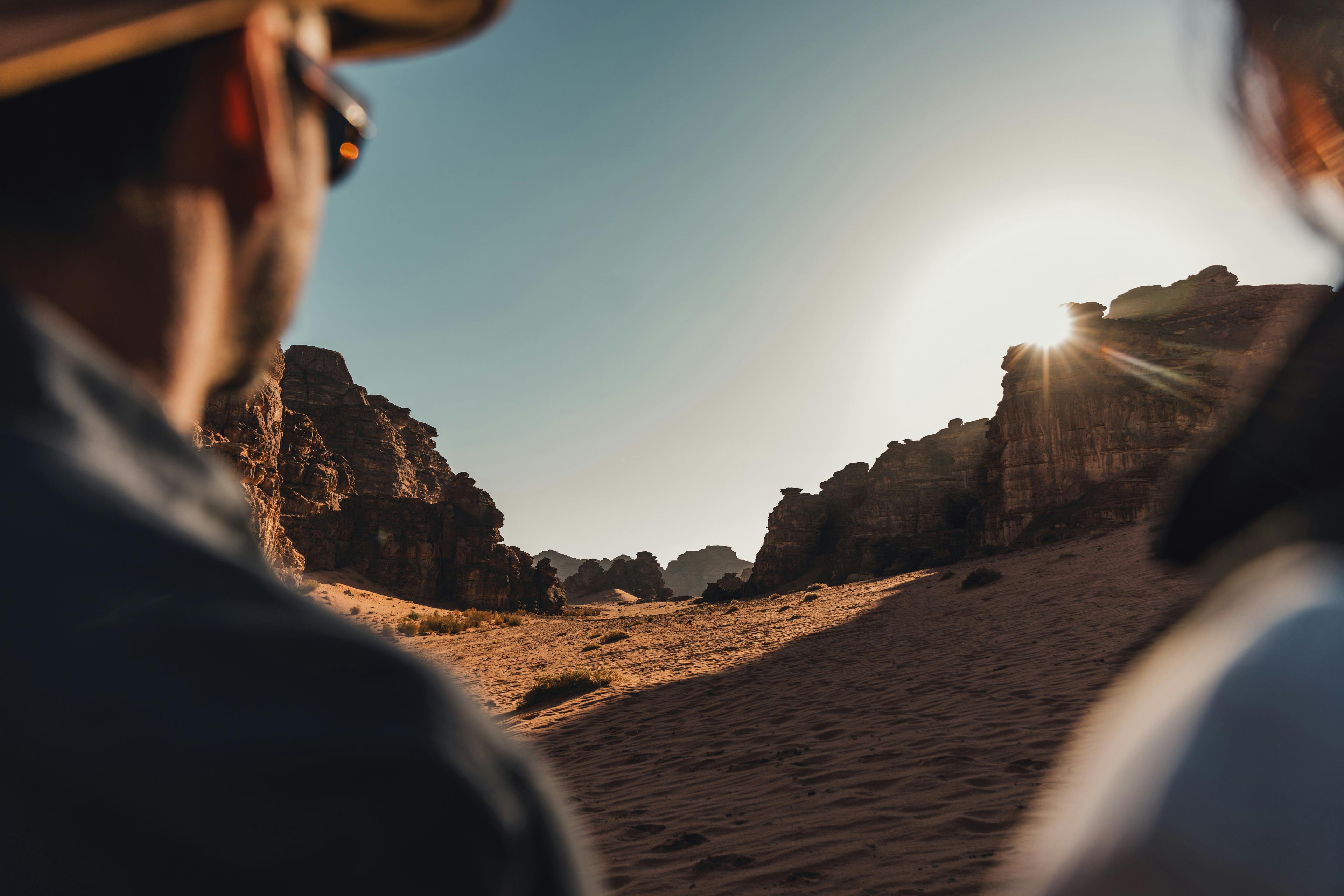 Two people observe a bright desert sunset illuminating rocky formations and sandy terrain.