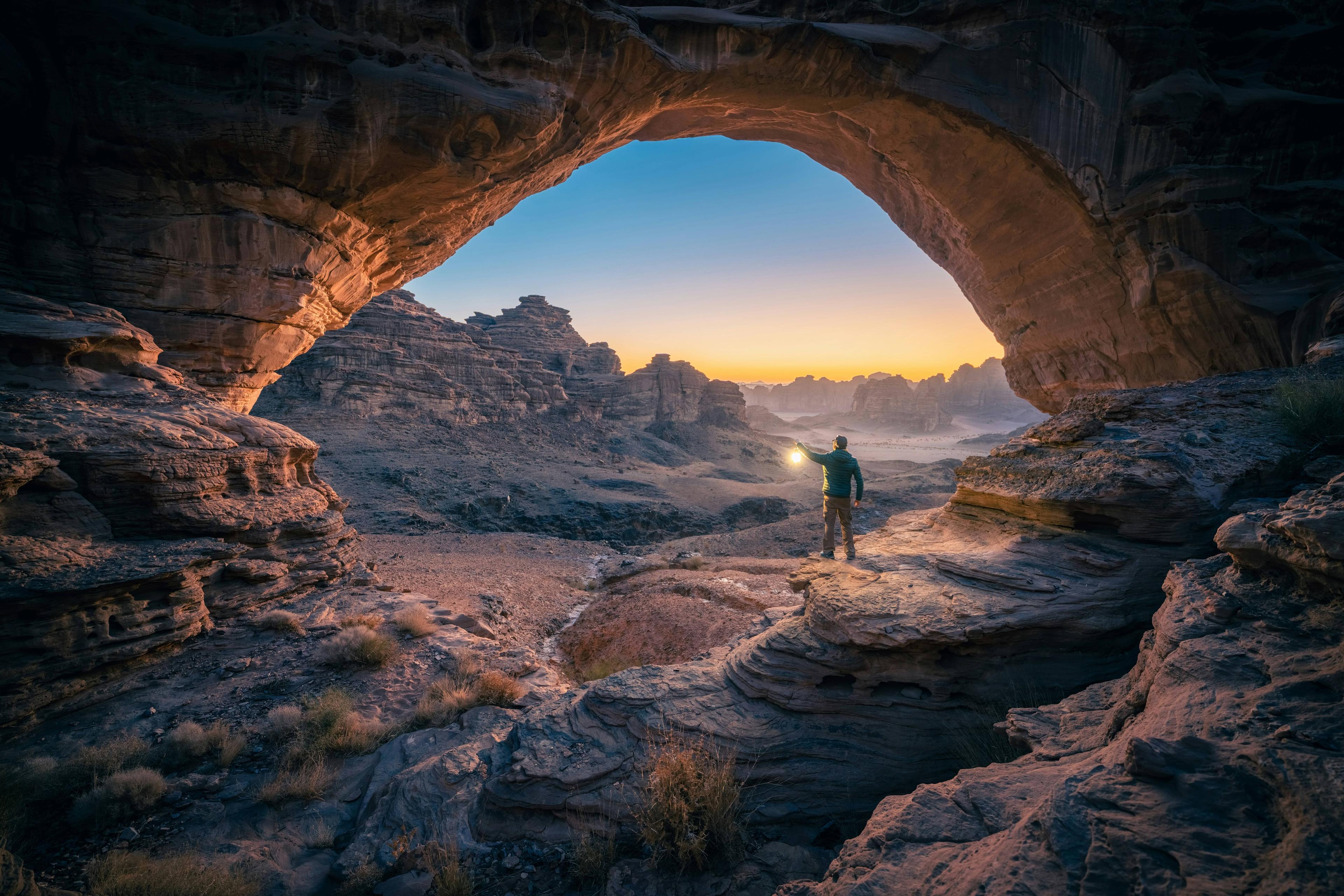 Person with a flashlight under a natural rock arch overlooks a vast desert at twilight.