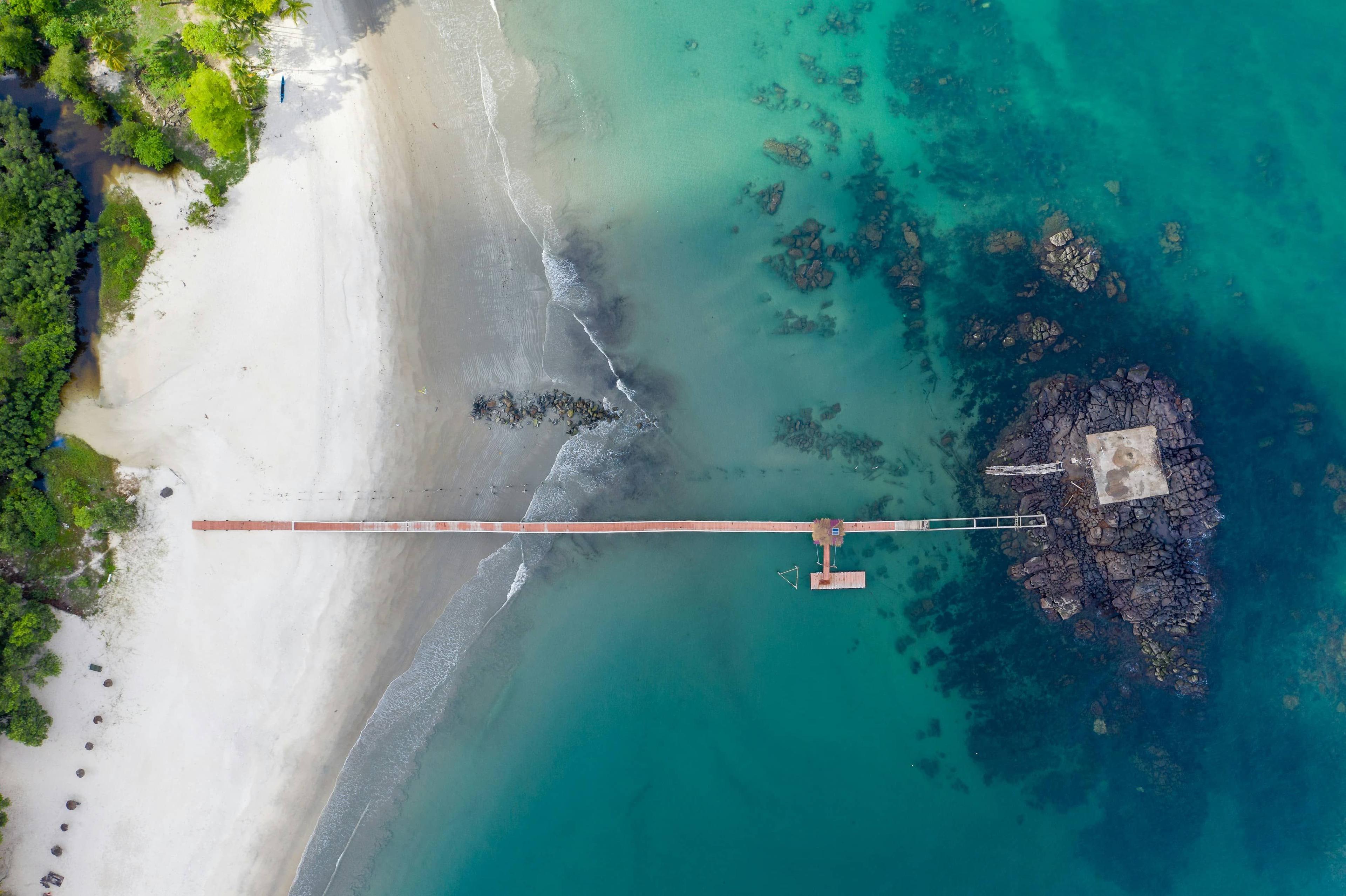 Aerial view: white sand beach, turquoise ocean, a pier connecting to a rocky island.